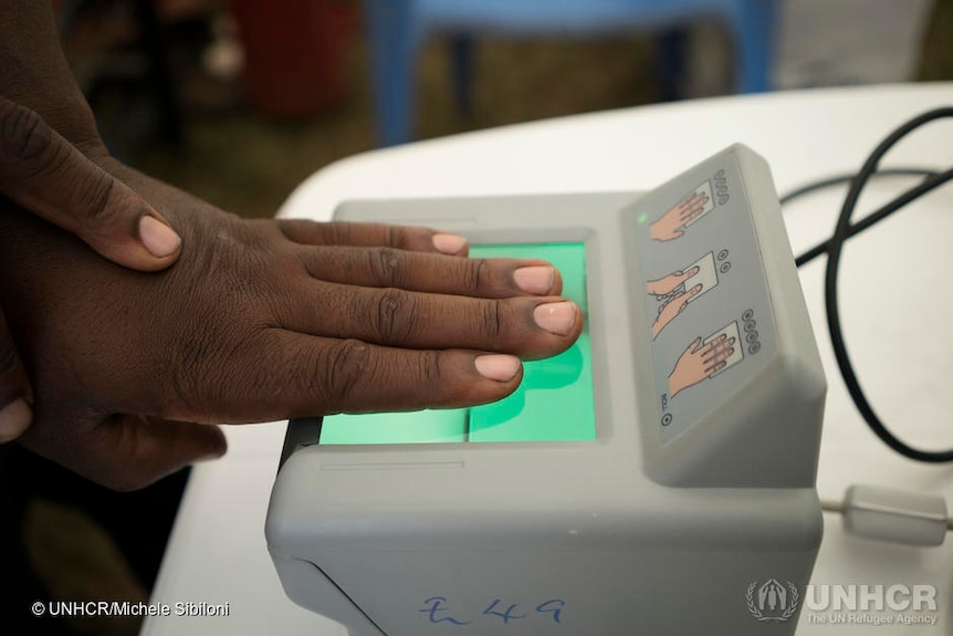 A man has his hand held on a machine, to take fingerprints.