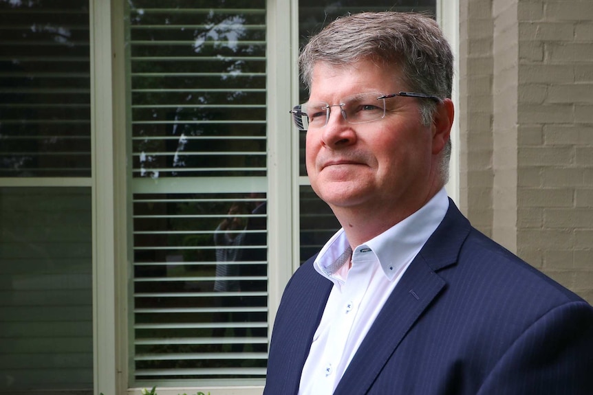 A bespectacled man with short grey hair stands outside, dressed in a dark suit.