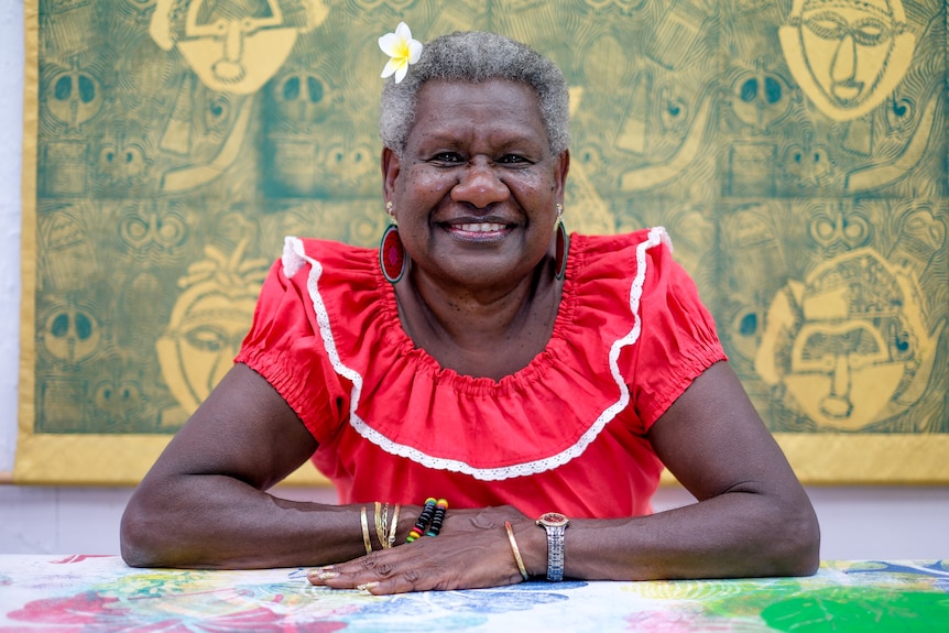 A woman wearing a red dress sits in front of a large screenprinted traditional Torres Strait Islander artwork