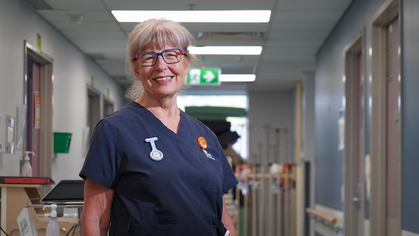 A grey haired woman standing in a hospital hallway smiles.