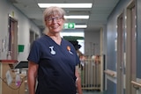 A grey haired woman standing in a hospital hallway smiles.