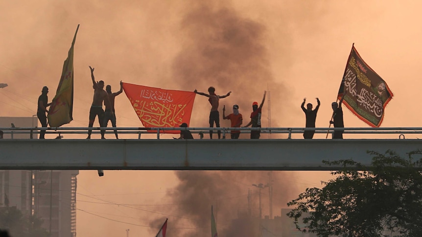 Anti-government protesters in Iraq hold flags on top of a bridge as smoke rises behind them.