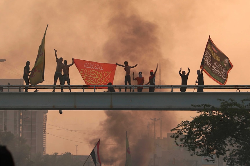 Anti-government protesters in Iraq hold flags on top of a bridge as smoke rises behind them.