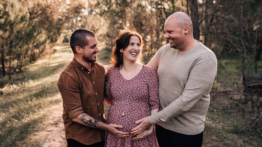 Posing for a photo on a tree-lined dirt track, two men press their hands against the belly of a pregnant woman who is smiling