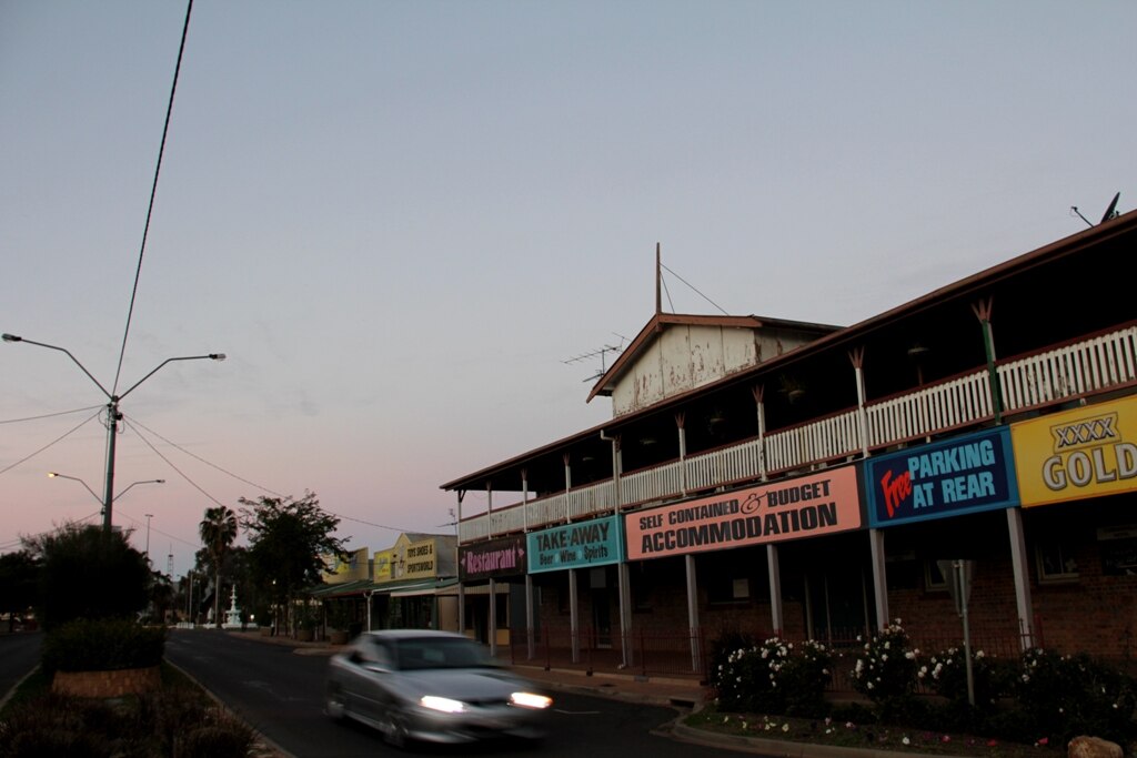 A car with headlights on heads along a quiet street in front of an old hotel.