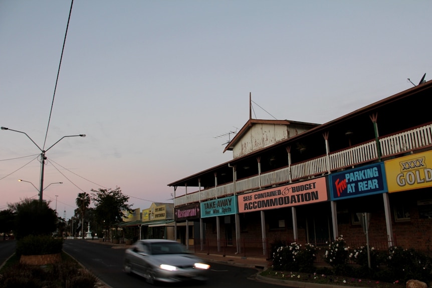 A car with headlights on heads along a quite street in front of an old hotel.