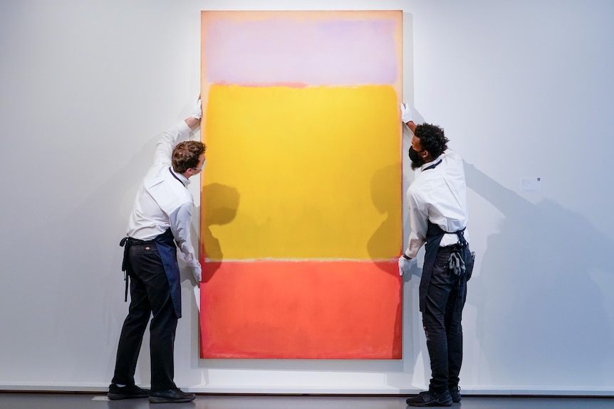 Two men in white shirts and black pants hand a tall red, green and blue painting on a white wall.