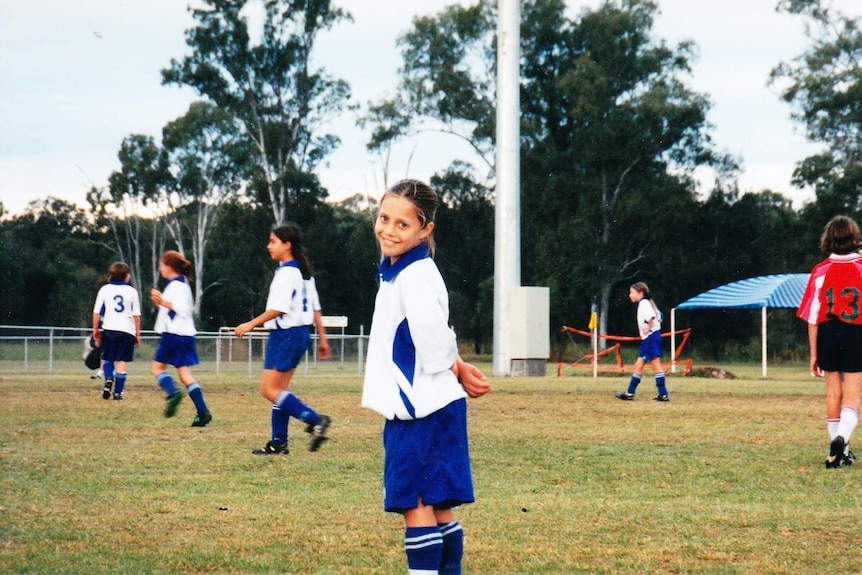 A girl dressed in blue and white football jersey and shorts stands on a soccer field during a game
