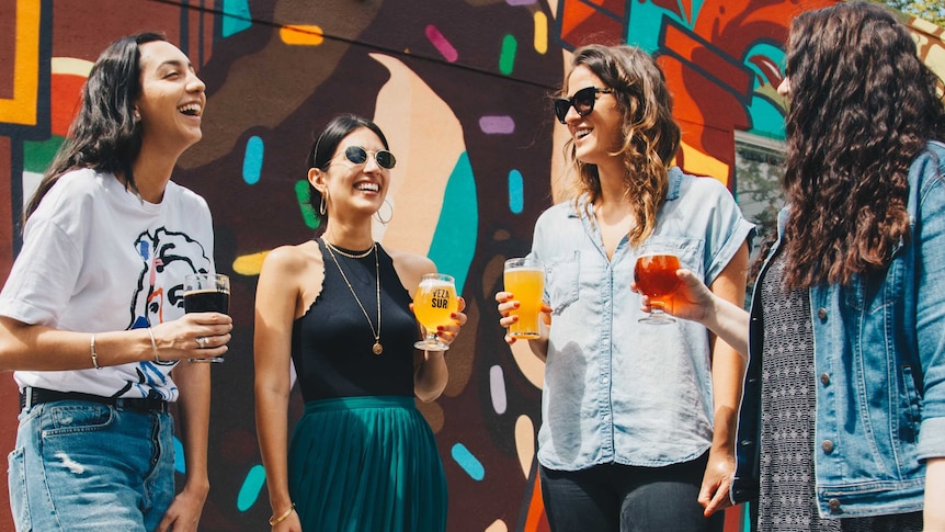 Four young women stand in front of a mural while holding beers