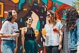 Four young women stand in front of a mural while holding beers
