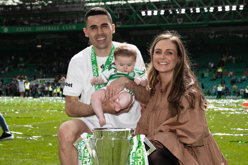 Celtic soccer player Tom Rogic smiles as he holds his daughter next to a woman, with a large trophy in front of them.