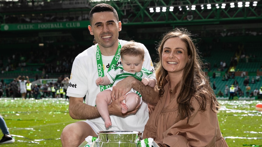 Celtic soccer player Tom Rogic smiles as he holds his daughter next to a woman, with a large trophy in front of them.