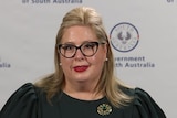 A woman with blonde hair, black glasses and a green top stands in front of a SA Government banner