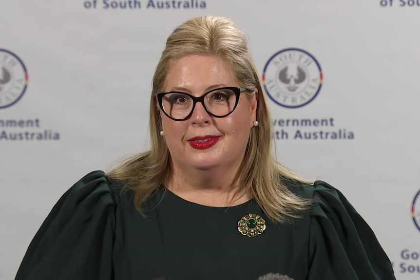 A woman with blonde hair, black glasses and a green top stands in front of a SA Government banner