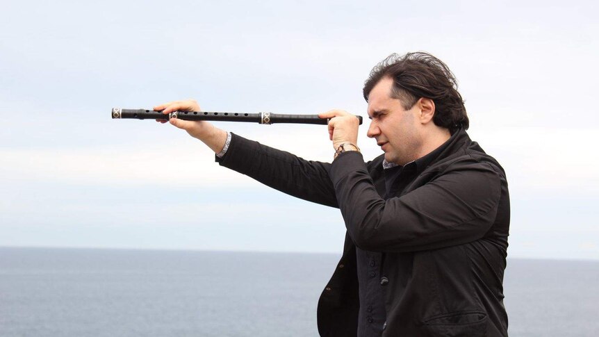 a member of the Balkan Ethno Orchestra looks through the body of a Bulgarian flute as if it's a telescope, in front of the sea