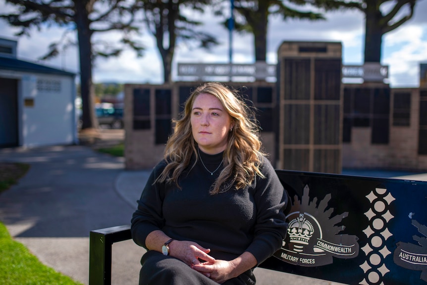 A woman in a black jumper looks contemplative while seated with columns and plaques of veterans' names behind her.