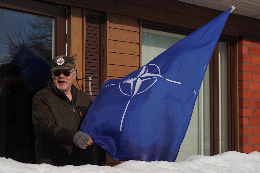 Retired Finnish combat engineer Ilkka Lansivaara poses with a NATO flag outside his home.
