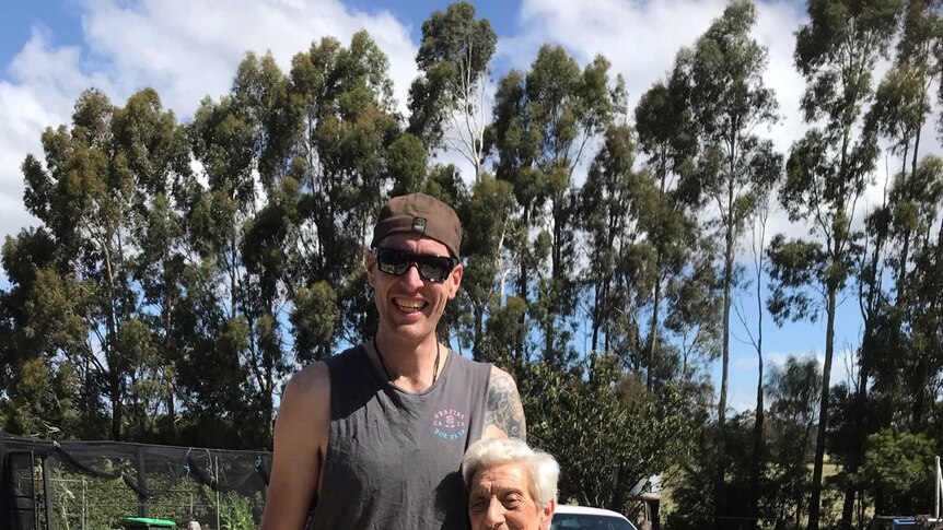 A tall man with tattoos and wearing a singlet, a backwards baseball cap and glasses with his arm around his grandmother