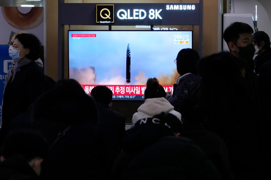 People watch a wall-mounted TV screen showing a missile launching into the air with Korean-language news tickers underneath.
