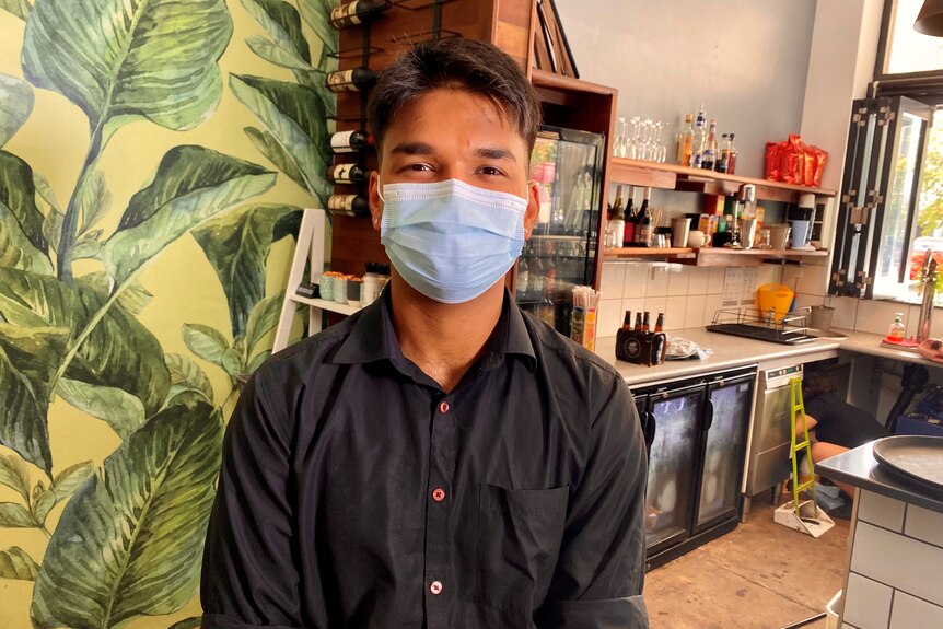 A man in a blue mask stands in a cafe, with tropical wallpaper and cafe kitchen benches behind him