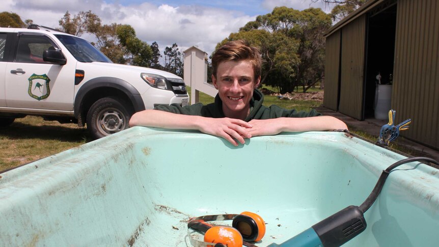 A dark-haired teenage boy with folded arms leaning over an old blue bath tub containing a sanding tool and orange ear-muffs.