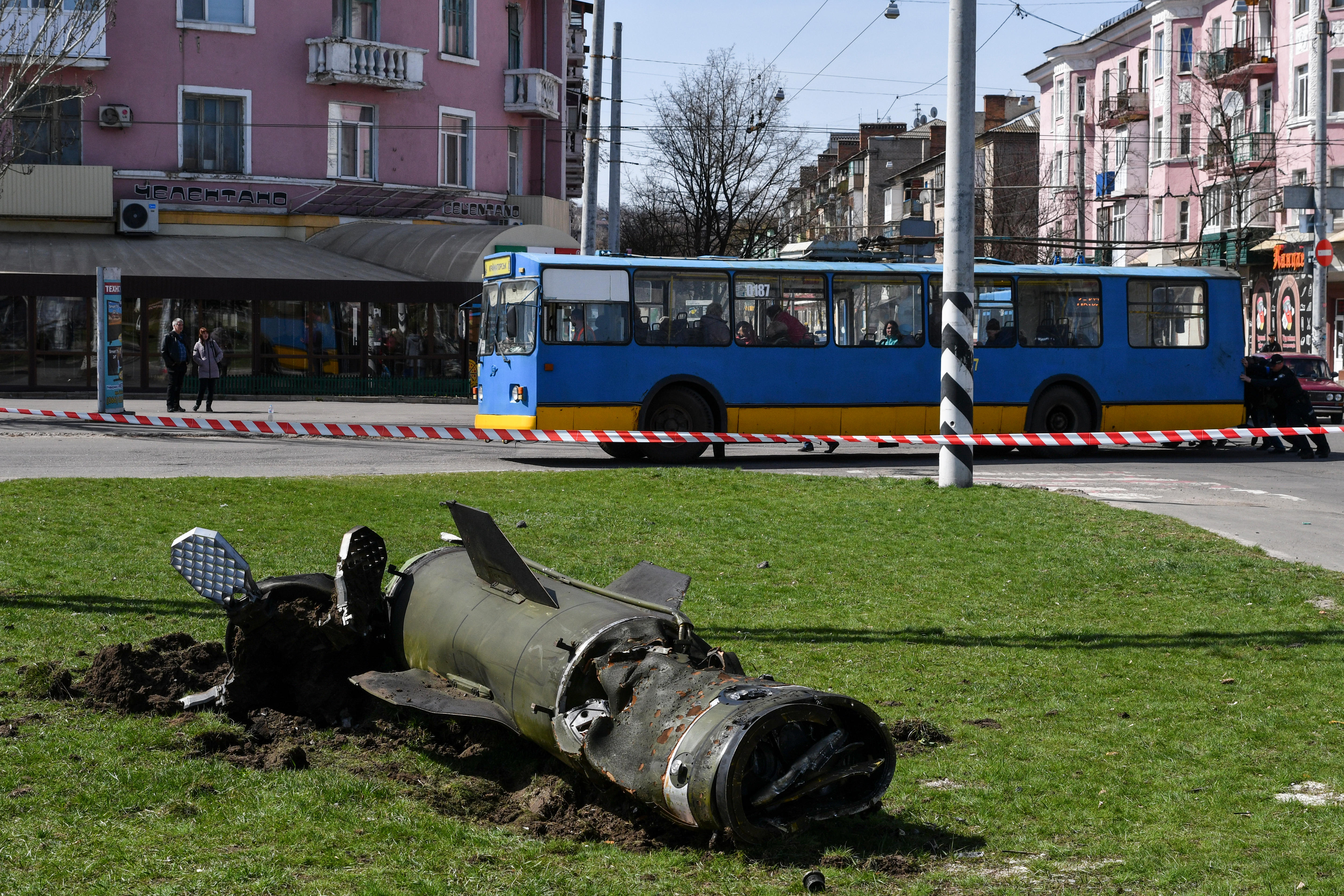 The remains of a rocket lie on a grass square with a bus and buildings in the background