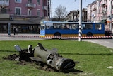 The remains of a rocket lie on a grass square with a bus and buildings in the background