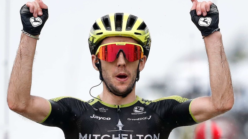 Simon Yates points both his index fingers to the sky while riding.