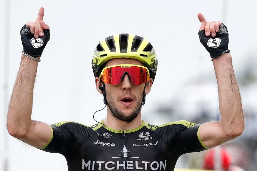 Simon Yates points both his index fingers to the sky while riding.