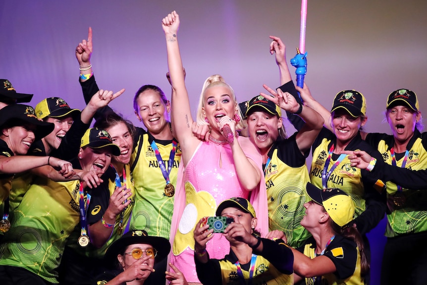 Katy Perry has one arm outstretched while singing into a microphone, with the Australian women's cricket team behind her.
