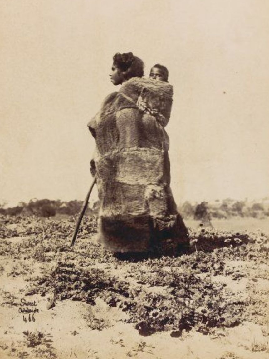 A woman wears a possum cloak with a baby on her back. Photo taken in South Australia, 1870s.