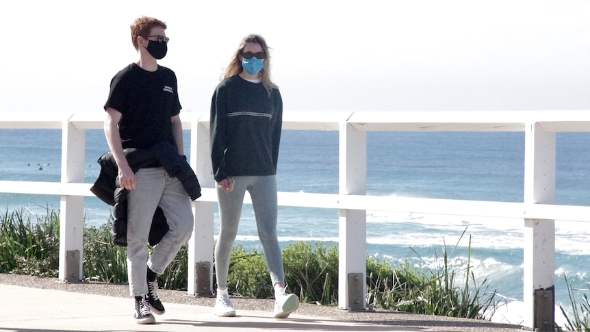 Two young people walk along the beack wearing face masks.