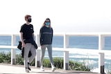 Two young people walk along the beack wearing face masks.