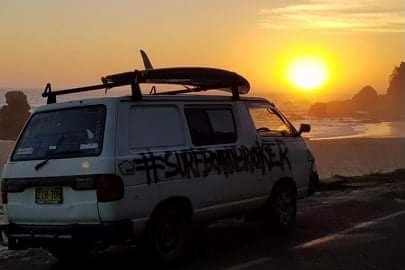 A van parked next to a beach at sunset, with a surfboard on the roof.