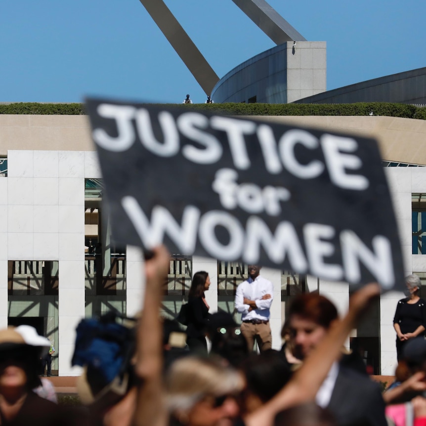 A protest sign reading JUSTICE FOR WOMEN is held up in front of Canberra Parliament House