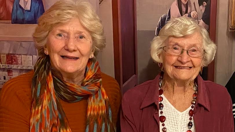 two women with light-coloured short curly hair sitting next to each other smiling