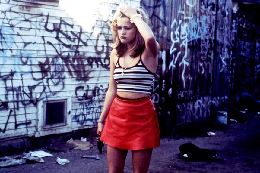 A young woman stands in red mini skirt with revolver, runs hand through blonde hair near graffitied derelict building and fence.