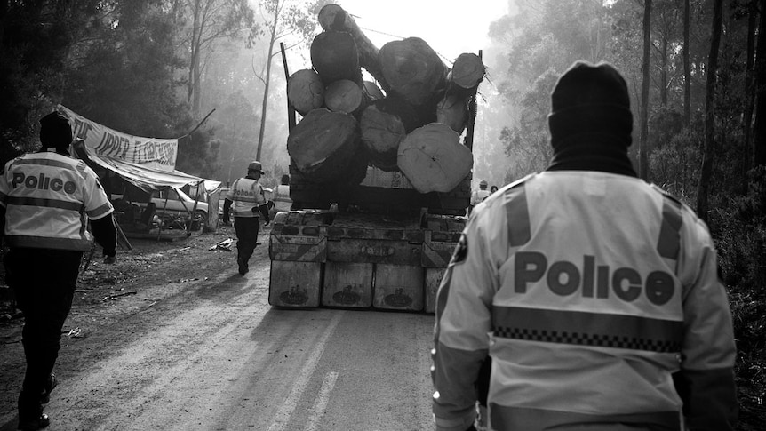 A laden log truck drives through the forest with police officers walking beside and behind it