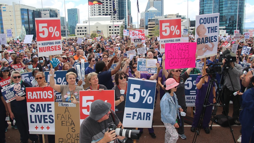 A wide shot of a crowd of nurses at a rally holding up signs and placards with tall CBD buildings behind them.