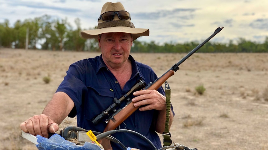 Brendan Slattery sits on a motorbike holding a gun, while wearing a battered wide-brimmed hat with sunglasses strapped to it.