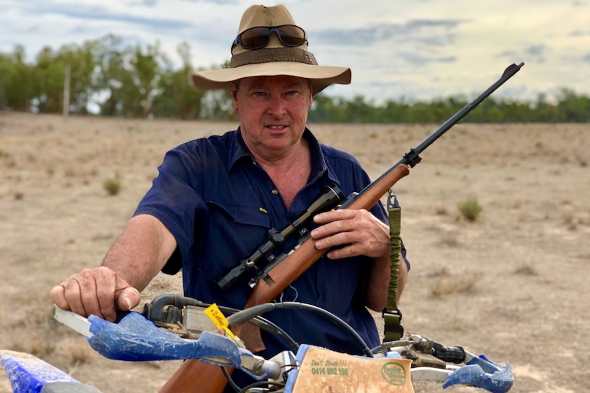 Brendan Slattery sits on a motorbike holding a gun, while wearing a battered wide-brimmed hat with sunglasses strapped to it.