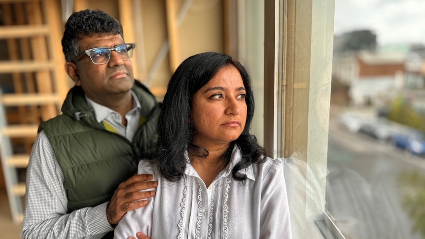 Siddarth stands behind Chetna with his hand on her shoulder as they both look out the window of their partially built apartment.