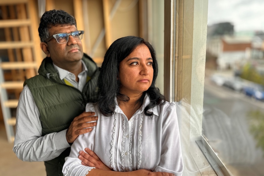 Siddarth stands behind Chetna with his hand on her shoulder as they both look out the window of their partially built apartment.