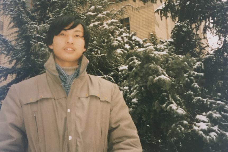 A young Chinese man in a trenchcoat with snow on the trees in the background, pictured in 1988 or 1989.