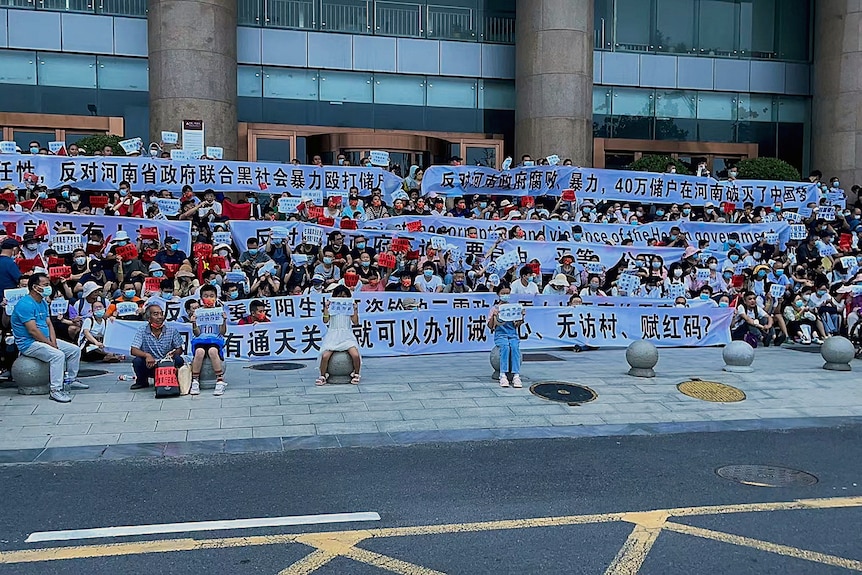 A group of hundreds of people sitting on the steps of a bank holding signs in Chinese