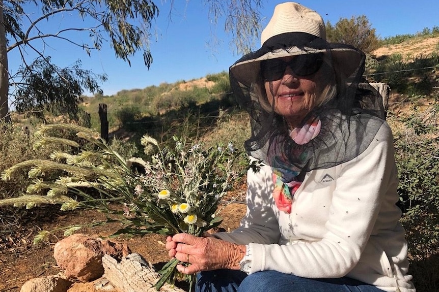 Nell Brook wears a hat with a flynet, holds some flowers while sitting in a garden in outback Queensland.