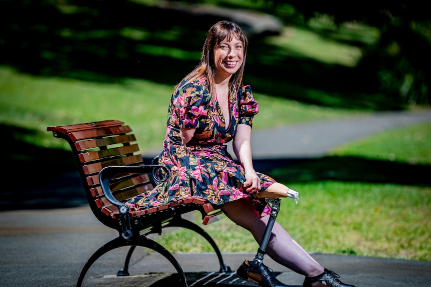 A smiling woman with a floral dress in the sunshine. She has a prosthetic leg and is sitting on a park bench