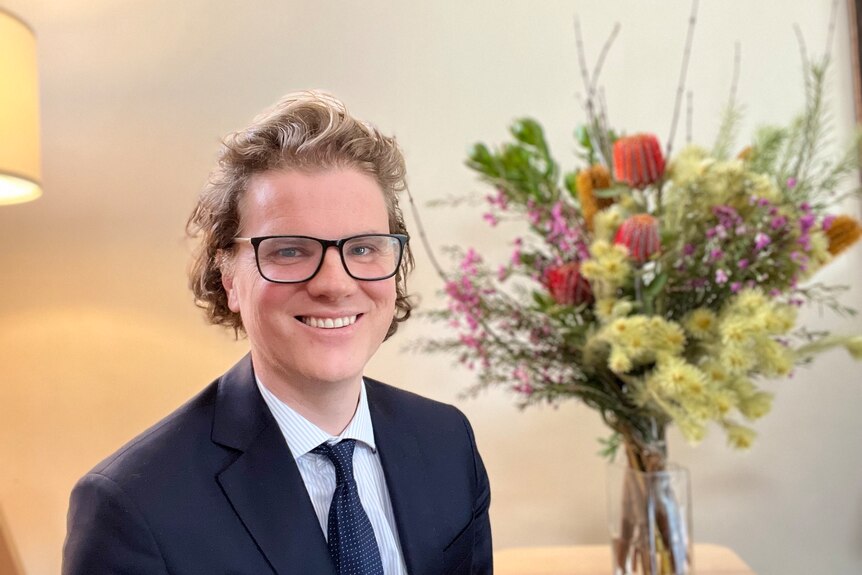 A smiling, bespectacled man with brushed back hair, wearing a dark suit and sitting next to a vase full of flowers.