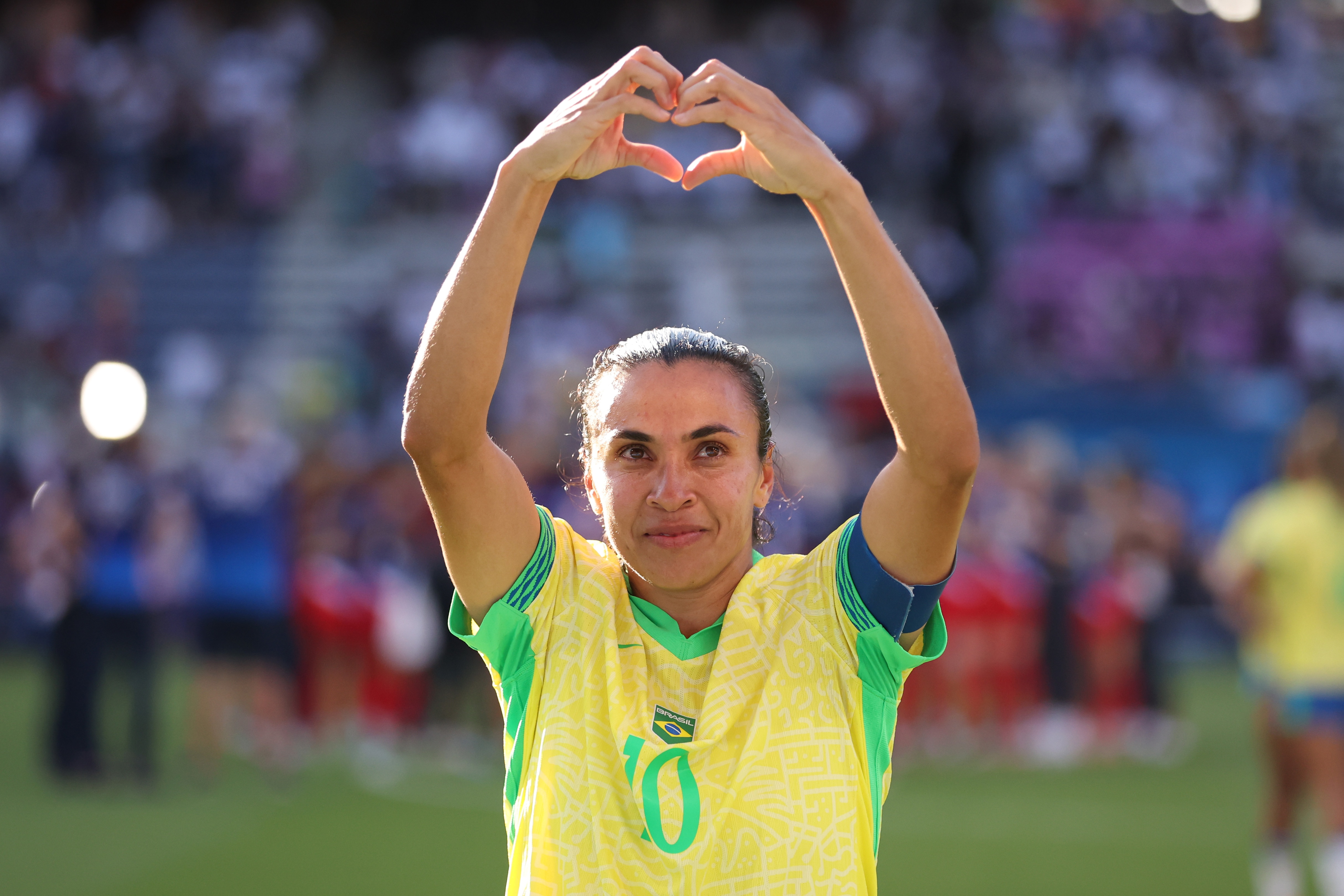 A soccer player wearing a yellow and green jersey with 10 on the front holds her hands in the shape of a love heart