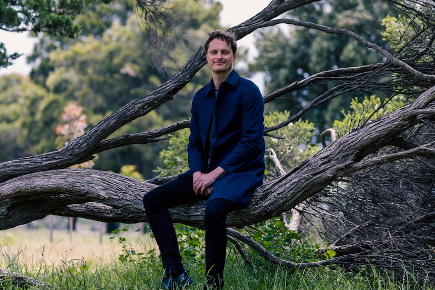 A man sits on a fallen tree branch in a park an smiles for a portrait